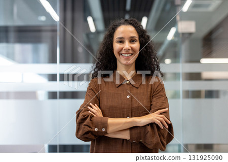 Professional businesswoman with curly hair smiling and looking at camera, standing with arms crossed in a modern corporate office, representing confidence and success Professional businesswoman with curly hair smiling and looking at camera, standing with arms crossed in a modern corporate office, representing confidence and success 131925090