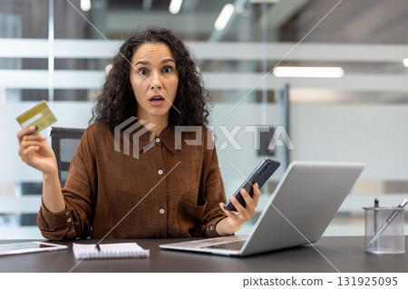 Woman holding a credit card and smartphone while looking shocked and stressed at a laptop screen in a modern office, experiencing financial problems, surprise, or unexpected debt 131925095