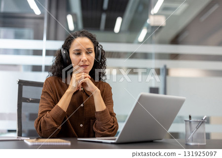 Focused woman wearing headphones, actively participating in a virtual conference or webinar from her desk, connecting remotely in a professional workspace 131925097