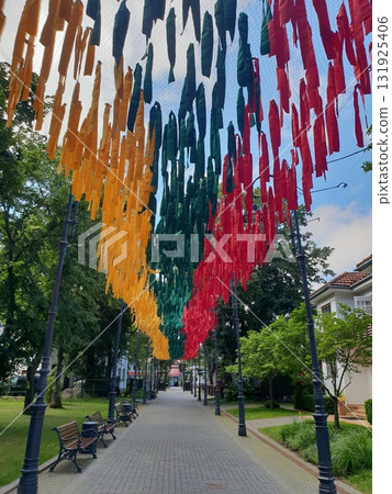 Colorful fabric decorations in vibrant hues of red, green, and yellow hang above a serene pathway lined with benches and lush greenery, creating a festive atmosphere 131925406