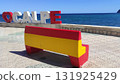 Colorful bench painted in red and yellow sits on a tiled promenade overlooking the blue sea, with large letters spelling a location in the background, creating a vibrant coastal scene 131925429