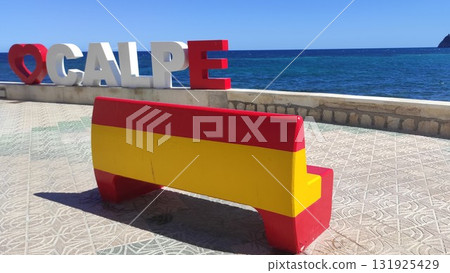 Colorful bench painted in red and yellow sits on a tiled promenade overlooking the blue sea, with large letters spelling a location in the background, creating a vibrant coastal scene 131925429