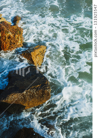 Waves crash against rugged rocks along the shoreline on a sunny day, illustrating the beauty of nature s coastal landscape Waves crash against rugged rocks along the shoreline on a sunny day, illustrating the beauty of nature s coastal landscape 131925547