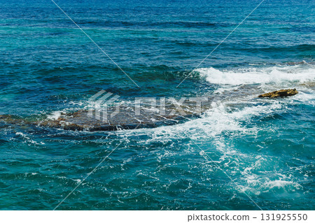 Waves crash against rocky shoreline under bright blue sky, capturing the tranquil beauty of the ocean at midday 131925550