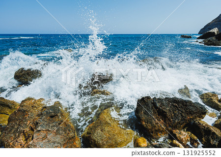 Waves crash against rocky shores at a sunny coastal spot, creating a spray of water that dances in the breeze under a clear blue sky Waves crash against rocky shores at a sunny coastal spot, creating a spray of water that dances in the breeze under a clear blue sky 131925552