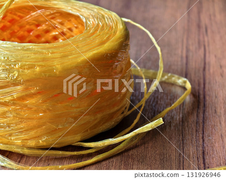 Close-up of yellow raffia plastic rope roll on wooden surface. Strong, durable, and flexible cord used for packaging, shipping, agriculture, DIY, and craft projects. 131926946