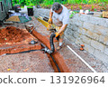 Man works diligently to connect drainage pipe while standing in trench beside house. 131926964