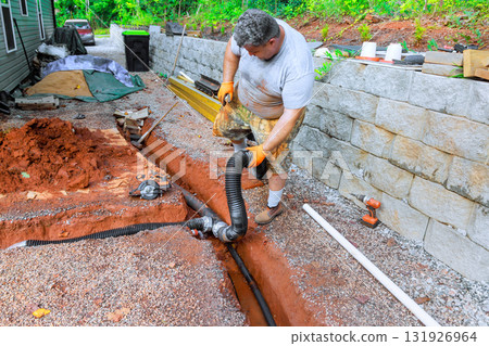 Man works diligently to connect drainage pipe while standing in trench beside house. Man works diligently to connect drainage pipe while standing in trench beside house. 131926964