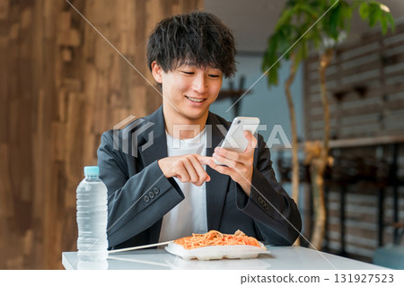 一位年輕的亞洲男性商人一邊吃盒飯，一邊在餐廳看著智慧型手機 131927523