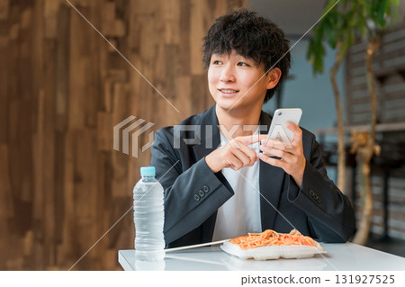 一位年輕的亞洲男性商人一邊吃盒飯，一邊在餐廳看著智慧型手機 131927525