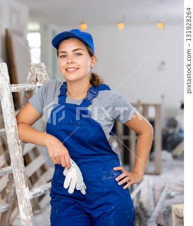 Portrait of contented young female builder in blue overalls during the repair of cottage Portrait of contented young female builder in blue overalls during the repair of cottage 131928264