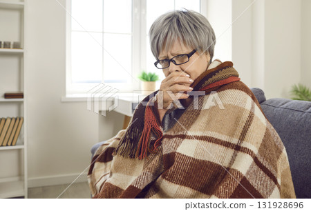 Sick upset senior woman wrapped in a blanket sitting on sofa at home with flu. Sick upset senior woman wrapped in a blanket sitting on sofa at home with flu. 131928696