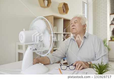 Senior man suffering from summer heat and trying to cool down by electric fan in his apartment Senior man suffering from summer heat and trying to cool down by electric fan in his apartment 131928717
