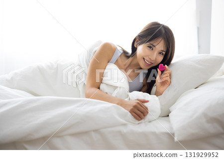 Smiling young woman holding a heart-shaped object in bed 131929952