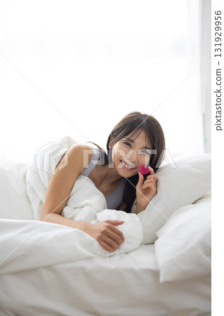 Smiling young woman holding a heart-shaped object in bed 131929956