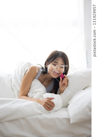 Smiling young woman holding a heart-shaped object in bed Smiling young woman holding a heart-shaped object in bed 131929957