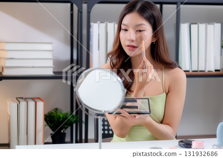 Happy young asian woman applying makeup with a brush on cheek looking mirror. Happy young asian woman applying makeup with a brush on cheek looking mirror. 131932686