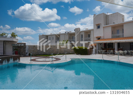 Modern hotel with a white facade, balconies, and a pristine pool surrounded by a tiled deck and shaded seating, set under a bright blue sky in Amalfi. 131934655