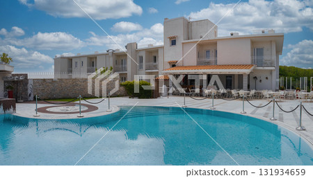 Modern hotel with white facade, terracotta roof, curved pool, outdoor seating, stone wall, greenery, and partly cloudy sky in Amalfi, Italy. Modern hotel with white facade, terracotta roof, curved pool, outdoor seating, stone wall, greenery, and partly cloudy sky in Amalfi, Italy. 131934659