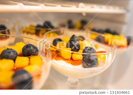 Elegant dessert glasses with creamy white base, mango chunks, blueberries, and red sauce, arranged on a refrigerator shelf for chilling. Elegant dessert glasses with creamy white base, mango chunks, blueberries, and red sauce, arranged on a refrigerator shelf for chilling. 131934661