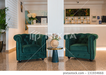 Luxurious hotel lobby featuring green velvet armchairs, a decorative table with flowers, a modern reception desk, and a greenery adorned wall in Amalfi. Luxurious hotel lobby featuring green velvet armchairs, a decorative table with flowers, a modern reception desk, and a greenery adorned wall in Amalfi. 131934668