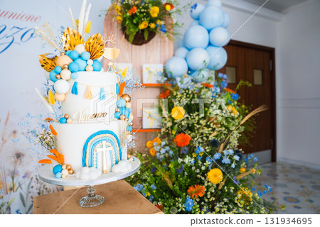 A multi tiered cake with blue, orange, and gold decorations, surrounded by floral arrangements and balloons, set in an indoor festive setting. 131934695