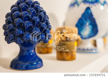 A vibrant blue ceramic pine cone ornament is displayed alongside jars of preserved food with burlap covers, evoking Amalfi's artisanal traditions. 131934731