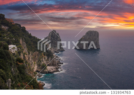 Faraglioni rocks rise from the sea near Capri, Italy, with cliffs lined by greenery and whitewashed buildings under a vibrant orange and pink sunset. Faraglioni rocks rise from the sea near Capri, Italy, with cliffs lined by greenery and whitewashed buildings under a vibrant orange and pink sunset. 131934763