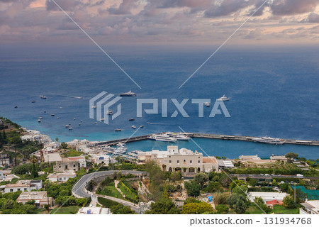 The image shows Capri's harbor with yachts in calm blue waters, whitewashed buildings, a winding road, lush greenery, and a partly cloudy sky. 131934768
