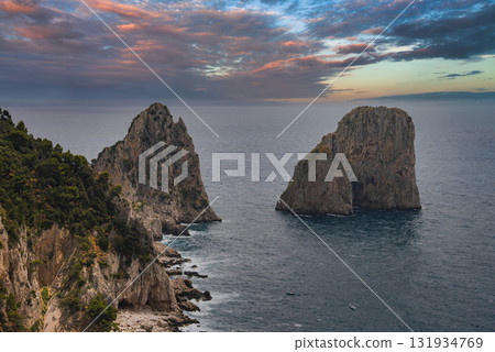 The Faraglioni rock formations rise from the Tyrrhenian Sea near Capri, Italy, with rugged cliffs and a vibrant sunset sky of orange, pink, and blue. 131934769