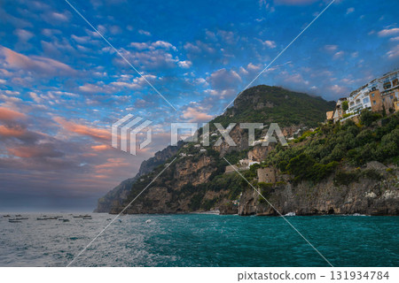 Positano, Italy, with pastel buildings on steep cliffs, lush greenery, and boats on turquoise waters under a colorful sunset sky. 131934784