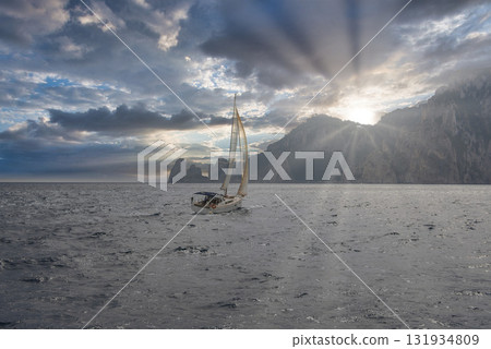 A sailboat glides on calm waters near Capri, Italy, with the Faraglioni rocks in the background. Sun rays pierce through clouds, illuminating the sea. 131934809