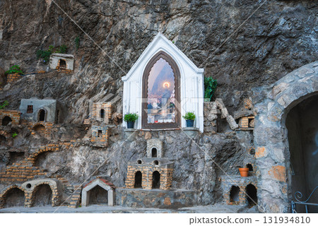 A carved rocky cliff features an arched frame with painted artwork, flanked by potted plants and miniature stone structures in Positano, Italy. 131934810