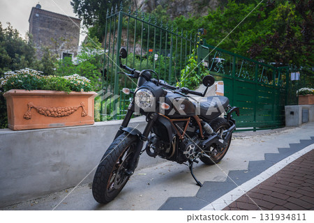 A black motorcycle is parked on a paved pathway by a green gate, with a terracotta planter, white flowers, and a stone building in Positano, Italy. 131934811