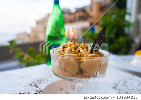 A creamy dessert in a glass dish topped with pistachios and chocolate, set against blurred terraced buildings and greenery in Positano, Italy. A creamy dessert in a glass dish topped with pistachios and chocolate, set against blurred terraced buildings and greenery in Positano, Italy. 131934815