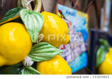 Vibrant ceramic lemons with green leaves are displayed in the foreground, with a colorful painting of a coastal town, likely Amalfi, in the background. 131934821