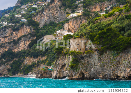 Positano, Italy features hillside buildings, a historic stone structure, rugged cliffs, lush greenery, and turquoise Mediterranean waters. 131934822