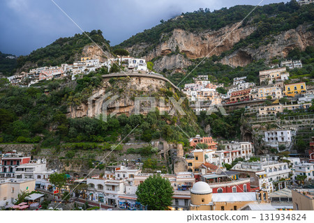View of Positano, Italy, with vibrant buildings on a steep hillside, a rocky outcrop with vegetation, and an overcast sky creating a dramatic scene. View of Positano, Italy, with vibrant buildings on a steep hillside, a rocky outcrop with vegetation, and an overcast sky creating a dramatic scene. 131934824