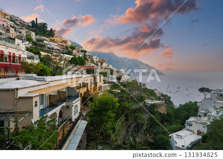 The town of Positano, Italy, features colorful terraced buildings on a hillside, with sunset skies, the Tyrrhenian Sea, boats, and lush greenery. 131934825