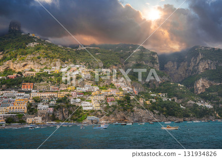 Positano on Italy's Amalfi Coast features vibrant buildings on a steep hillside, turquoise waters with boats, rugged cliffs, and dramatic skies. 131934826