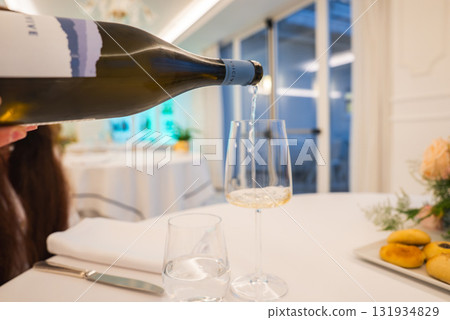 A bottle of white wine is poured into a glass on a table with white linens, pastries, and a floral centerpiece in a bright Amalfi restaurant interior. A bottle of white wine is poured into a glass on a table with white linens, pastries, and a floral centerpiece in a bright Amalfi restaurant interior. 131934829