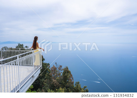 A beautiful woman in a yellow dress stands on a white balcony above lush greenery, overlooking the Tyrrhenian Sea with boats and distant landforms in view. A beautiful woman in a yellow dress stands on a white balcony above lush greenery, overlooking the Tyrrhenian Sea with boats and distant landforms in view. 131934832