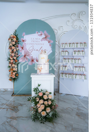 Elegant first communion setup with a pastel green backdrop, floral cake on a white pedestal, peach roses, party favors, and ornate white wall detailing. Elegant first communion setup with a pastel green backdrop, floral cake on a white pedestal, peach roses, party favors, and ornate white wall detailing. 131934837