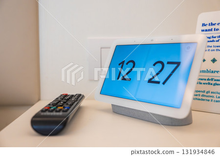 Modern digital clock displaying 12 27 placed on a white surface next to a black remote control. Background includes a printed sign and wall socket. Modern digital clock displaying 12 27 placed on a white surface next to a black remote control. Background includes a printed sign and wall socket. 131934846