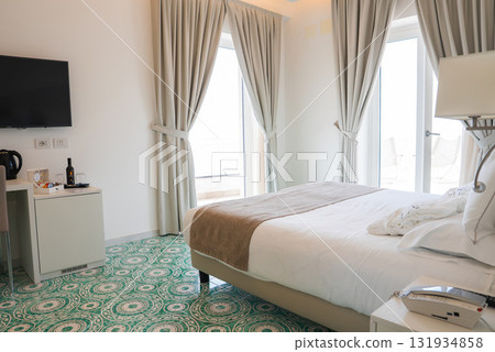 Bright hotel room in Amalfi, Italy, featuring white linens, tiled flooring, a desk with wine, large windows, and a glimpse of an outdoor balcony. 131934858
