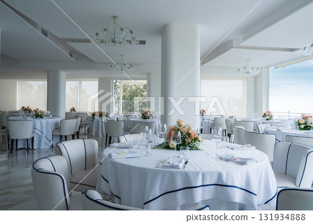 Dining area with round tables, white tablecloths, floral centerpieces, and large windows offering natural light and partial views of greenery and sky. 131934888