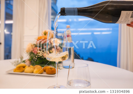 A glass of white wine is poured on a table with bread rolls and flowers. Large windows reveal the Tyrrhenian Sea and coastal cliffs in Amalfi, Italy. 131934896