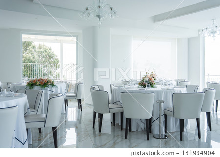 Modern dining space with white walls, marble floors, round tables, floral centerpieces, and large windows offering partial views of greenery and sea. 131934904