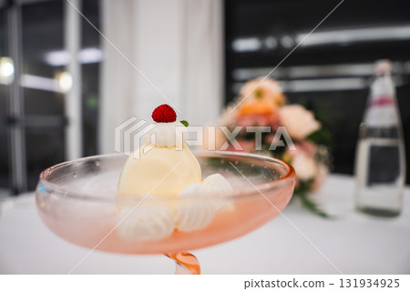 A creamy yellow dessert in a pink glass, topped with whipped cream, a red berry, and mint. Blurred floral arrangement and water bottle in background. A creamy yellow dessert in a pink glass, topped with whipped cream, a red berry, and mint. Blurred floral arrangement and water bottle in background. 131934925