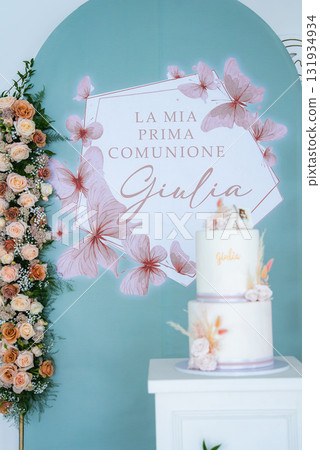 Decorative setup for a first communion with a pastel green backdrop, floral illustrations, a two tiered white cake, and a rose arrangement. Decorative setup for a first communion with a pastel green backdrop, floral illustrations, a two tiered white cake, and a rose arrangement. 131934934
